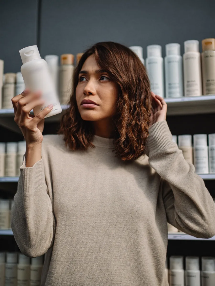 Mulher olhando em dúvida para um frasco de shampoo diante de uma prateleira cheia de produtos